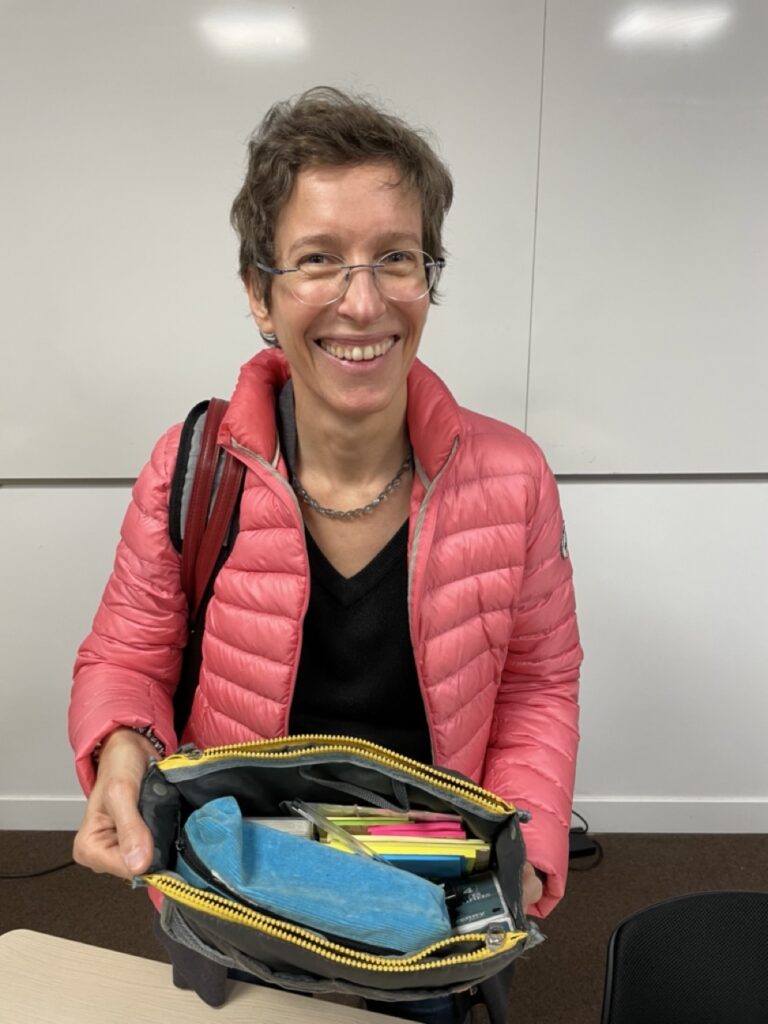 A smiling woman holds an open bag. Inside the bag, the kit of an agile coach and trainer: post-it notes, kit, pens...